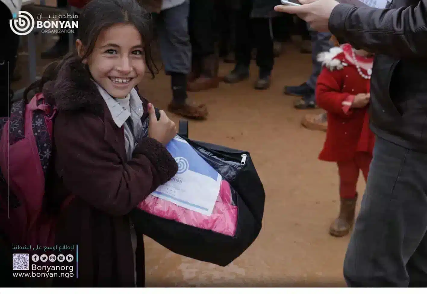  A smiling girl holds a bag of food and clothing in front of a crowd of people. The bag has the Bonyan Organization logo on it.